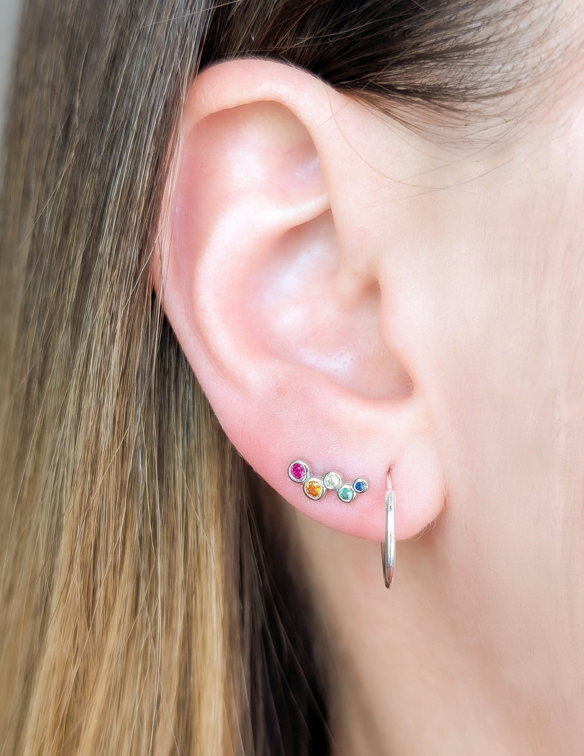 Boucle d’oreille Luma à zircons colorés portée au lobe, à côté d’un petit anneau argenté.